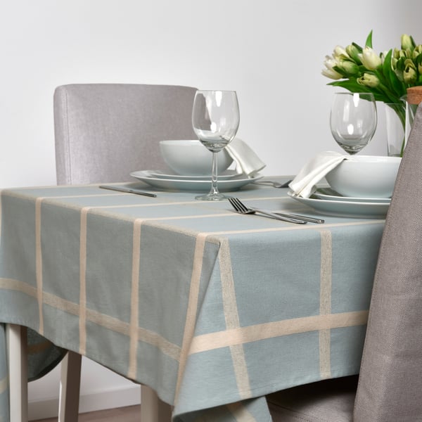 Dining table set with blue plaid tablecloth, plates, glasses, utensils, and white flowers in vase.