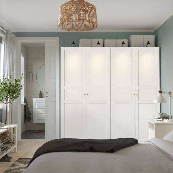Modern bedroom with tall 6-door white PAX wardrobe, gold handles, top white boxes, pendant lamp, partial bathroom view, clean aesthetic.