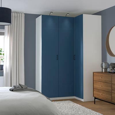 Large blue PAX wardrobe with white frames, modern design, stands tall beside wooden drawers and bed, sleek handles, vertical stripes.