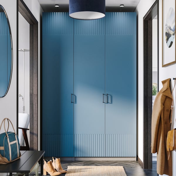 Modern blue PAX wardrobe dominates room, tall with sleek design. Coats hang nearby, bags and boots below, mirrored door reflects space.