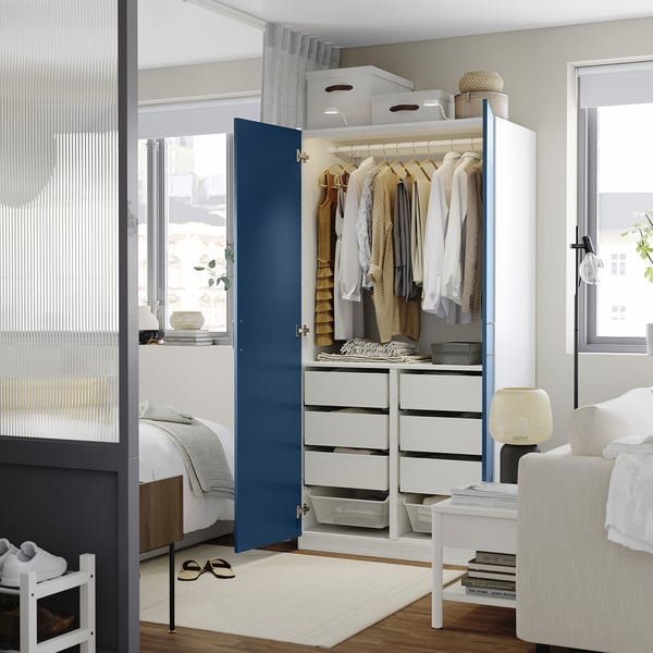 A modern open wardrobe displaying clothes on hangers, with four drawers below, set in a bright room with a large window.