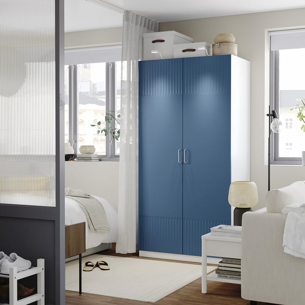 Modern bedroom with blue PAX wardrobe & white chair. Wardrobe tall, wide, with silver handles. Large window beside it.