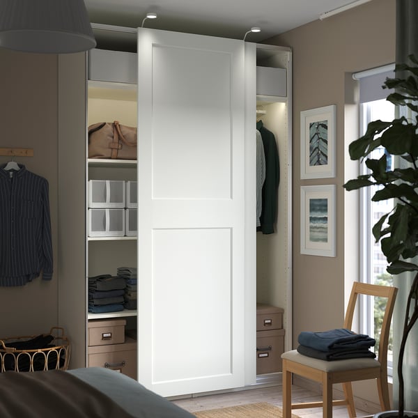 Modern bedroom with white PAX wardrobe, sliding doors, wood accents, hanging clothes, shelves, and houseplants.