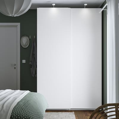 Minimalist bedroom with large PAX wardrobe, sleek doors, and lighting. Grey door, green bed cover, white curtains, hat, and towel by door.