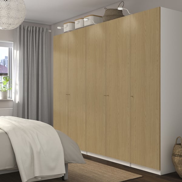 Large light-colored wooden closet with multiple doors next to a bed; white comforter and grey curtains visible.