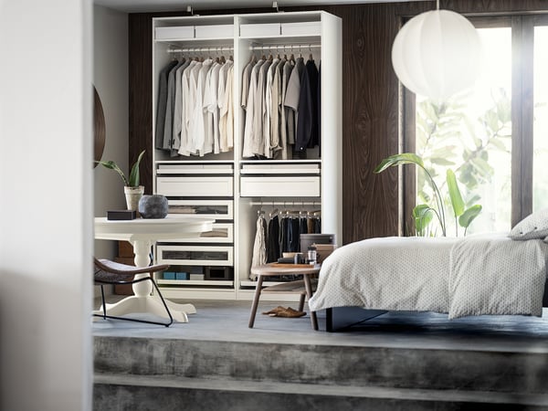A modern bedroom featuring a white PAX wardrobe with hanging clothes, drawers, and glass shelves. It includes a striped bench and a polka-dot bed.