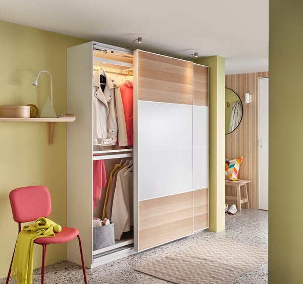 Modern bedroom with open wooden PAX wardrobe, light-colored clothes, red chair, yellow accents.