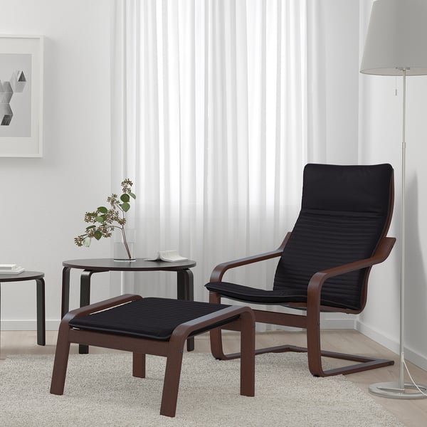 Modern living room with black & brown poang armchair, footstool, and small table. Armchair and stools curve design, white curtains, grey carpet.