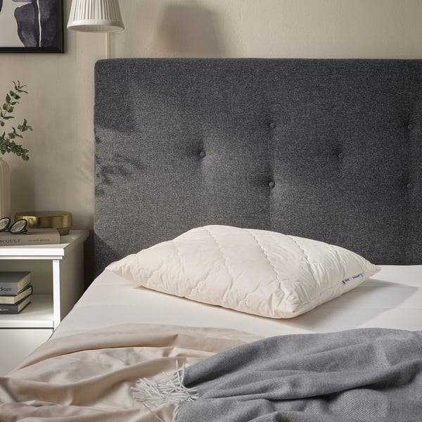 Grey tufted headboard with white RÅDHUSVIN pillow, light linens, bedside table with books, lamp, and plant.