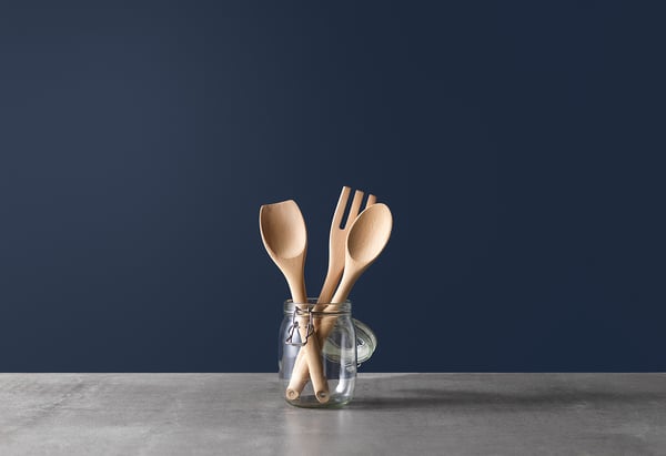 Three wooden spoons in clear glass jar on grey table, blue wall background.