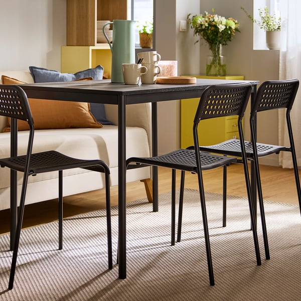 Compact dining set in cosy room. Black table with metal frame, wooden top, four chairs, and green pitchers. Perfect for small spaces.