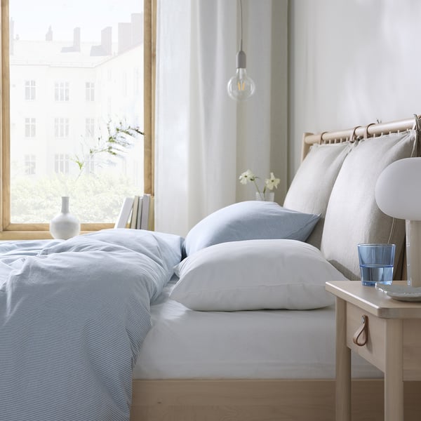 Bright bedroom: white bedding, wooden bed, hanging bulb, window with city view.