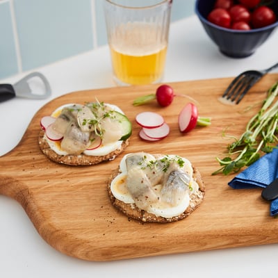Wooden board holds two open sandwiches topped with herring, sliced vegetables, and greens. Glass of yellow drink, knife, and radishes nearby.