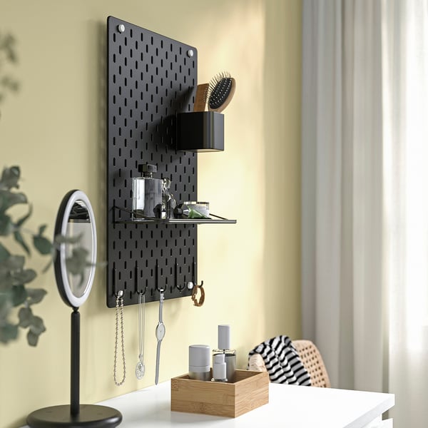 Black SKÅDIS pegboard with hooks, shelves, and compartments holding beauty items, jewellery, and plants on a yellow wall.