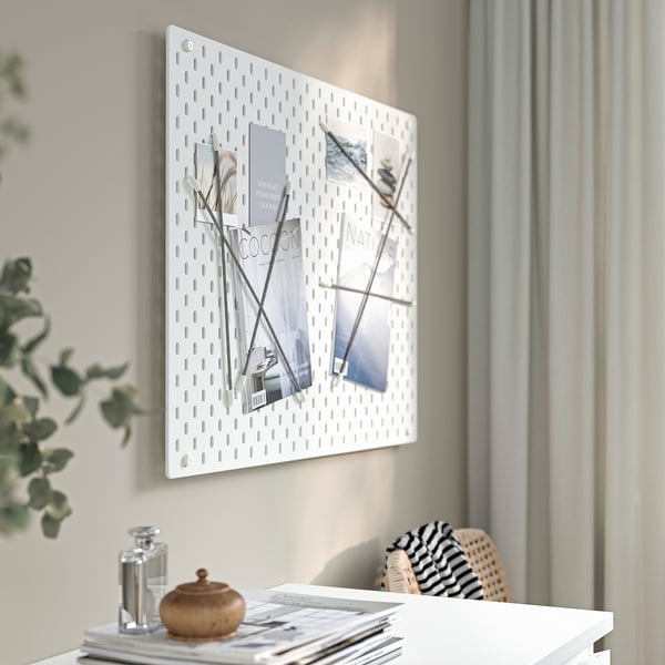 A white wall-mounted SKÅDIS pegboard displays organised photos and magazines. A stylish desk below holds a decorative plant, a jar, and a woven bag.