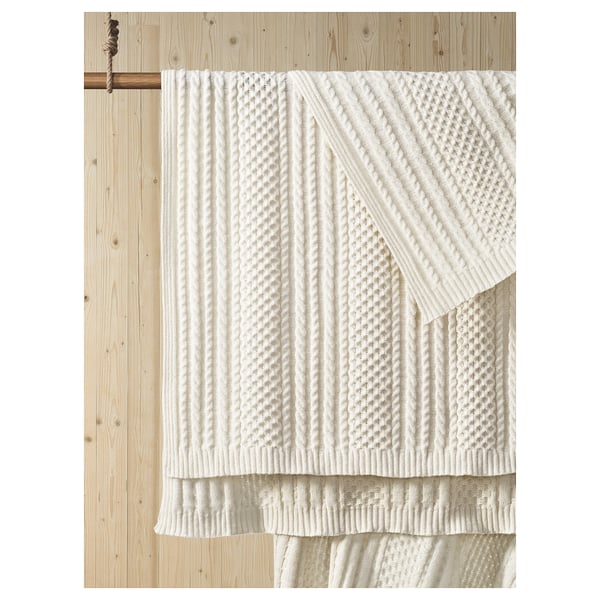 White knitted blanket on wooden wall. Soft, textured, and machine-washable. Made from 100% recycled polyester.