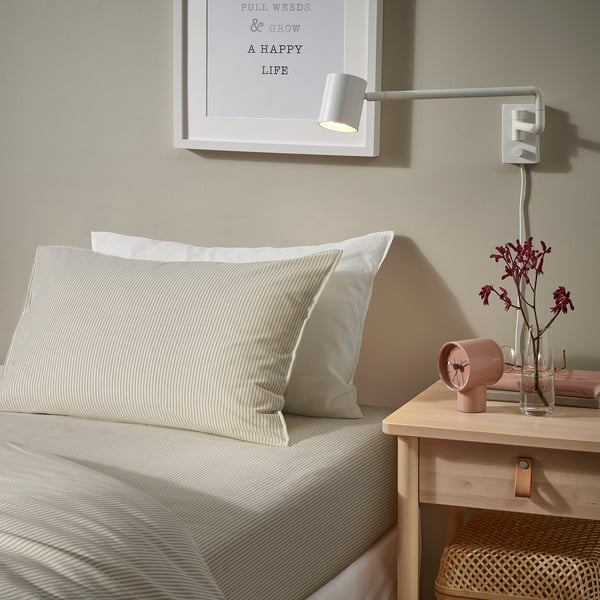 Bed with SOLFIBBLA white striped pillows, wall light, small bedside table with vase, clock & book.