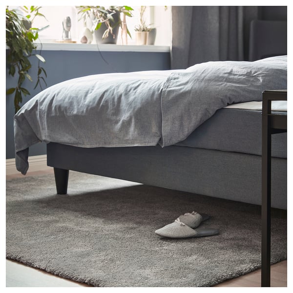 Bed with grey bedding, black legs, grey slippers, and a grey rug on a wooden floor.