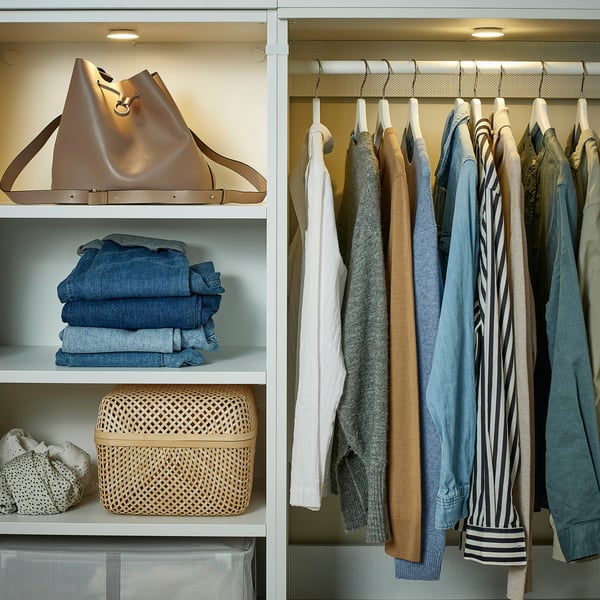 Organised closet with clothes and accessories. Spotlight illuminates shelves featuring a large handbag, folded jeans, and a woven basket.