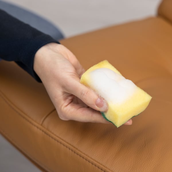 A hand holds a yellow sponge cleaning a brown leather surface, demonstrating TRIXIG leather cleaners ability to clean and maintain leather furniture.