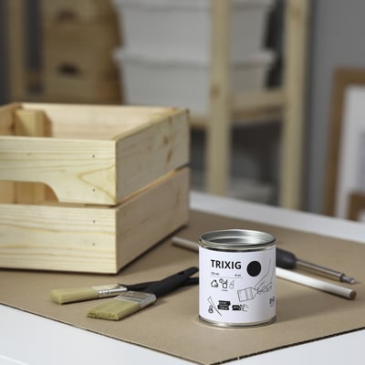 Small white can of TRIXIG paint sits on cardboard, surrounded by brushes and an empty wooden box, ideal for small diy projects.
