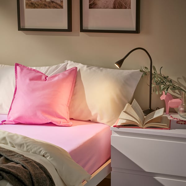 Bed: VÅRVIAL pink cotton sheet, fits 26cm mattress, with white pillows, beside black lamp & book.