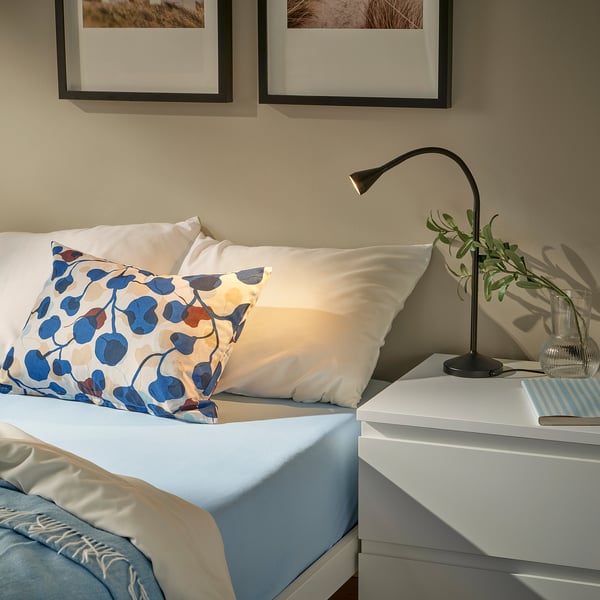 Blue VÅRVIAL pillow on bed, with white pillows and black lamp. Abstract blue-red pattern.