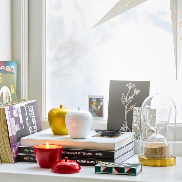 Shelf by window with apple candle, jars, hourglass, books, plant art.