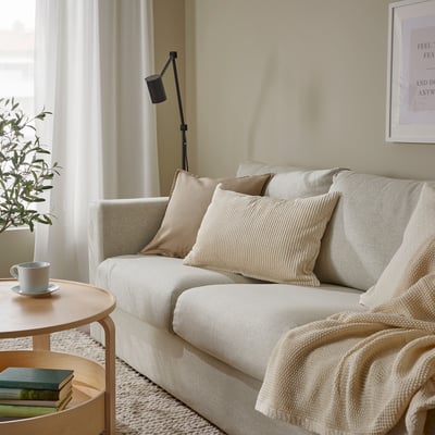 Rahulik elutuba kreemika ÅSVEIG diivani, tekstuursete patjade, puidust lisalauaga, kus on kruus ja raamatud, taim ja põrandalamp.