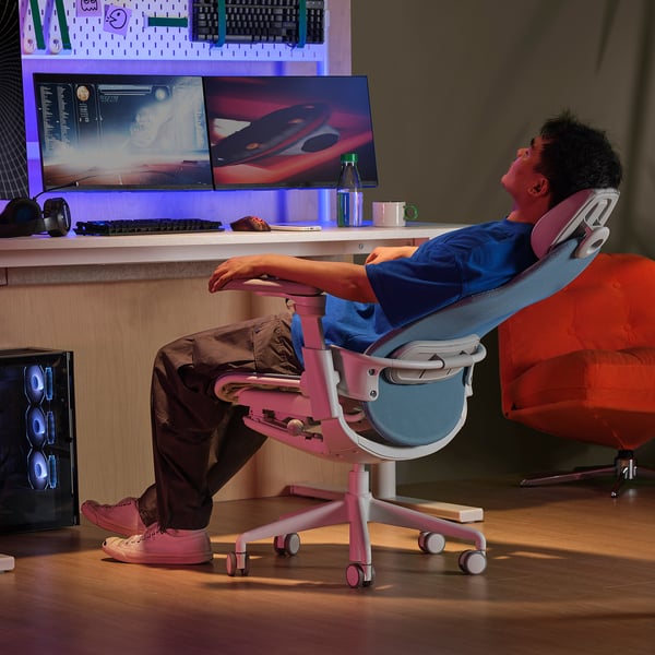 Mängija lamab roosas BÄSTBOLL toolis, jalad püsti, nautides ergonoomilist mugavust ja võrgust seljatuge koos kahe monitori ja varustusega.