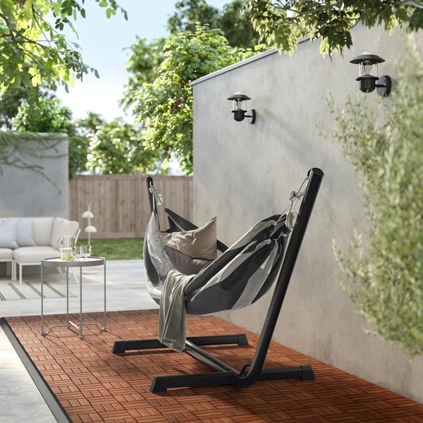 Kaasaegne õuevõrkkiigetool musta aluse, halli kanga, patjadega, puidust tekil, külglaua ja seinavalgustite kõrval.