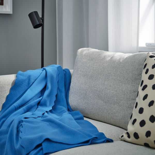 Hubane sinine DAGGKÅPOR fliisist pleed, mis katab valdavalt helehalli diivanit, läheduses mustad täpilised padjad ja lamp.