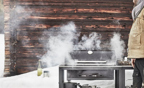 Inimene grillib õues musta GRILLSKÄR grilliga, mis eraldab suitsu. Lumi katab maad ja taustal on puidust majake.
