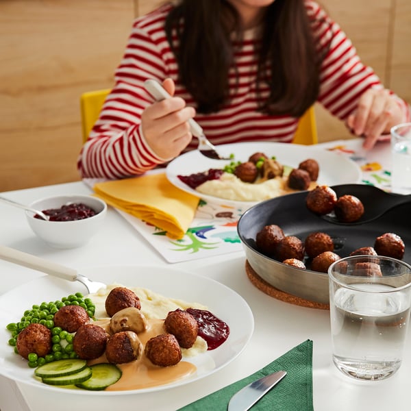 Triibulises särgis inimene, kes sööb taldrikult lihapallide, kartulipüree, herneste ja kurkidega valgel laual koos klaasi veega.