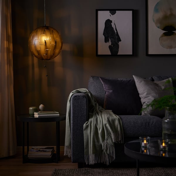 Hubane elutuba IKEA PS 2014 lambiga pimeda diivani kohal, mis heidab sooja valgust. Lisalaual on raamatud ja sisekujundus, kunst seinal.