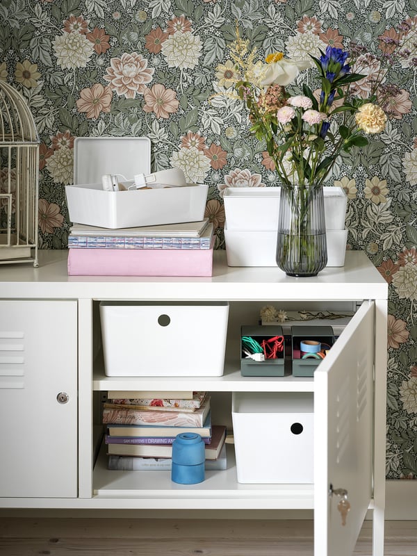 Valge IKEA PS metallkapp; lilleline tapeet; ülaosa: hoiukastid, raamatud, vaas lilledega; alumine: avatud uks, mis paljastab raamatud, konteinerid.