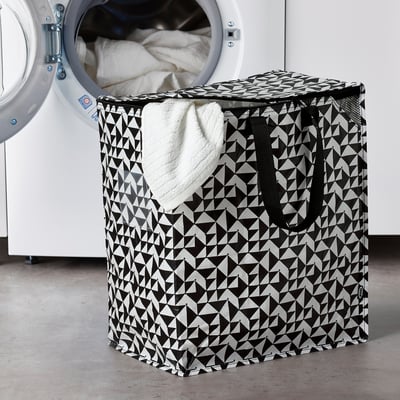 KNALLA mustvalge geomeetriline pesukorv mahutab palju esemeid, mis on paigutatud suurema valge pesumasina kõrvale.