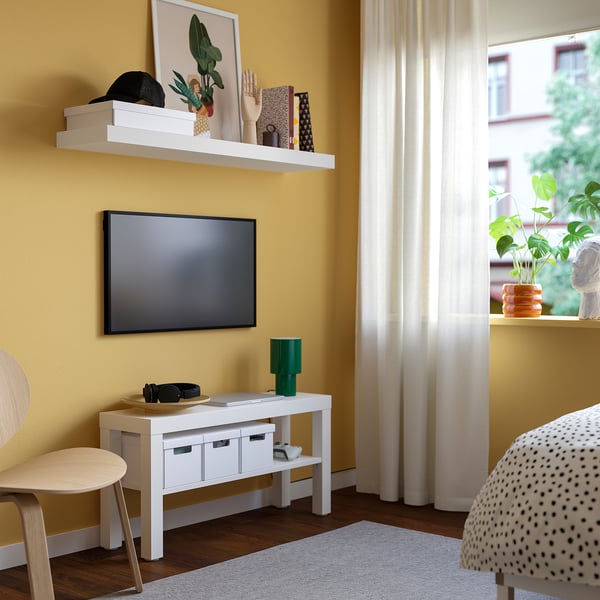 Kaasaegne kollaste seintega elutuba, valge televiisor, lameekraaniga televiisor, roheline lamp, puidust tool, täpiline tugitool, suur kardin ja taim.