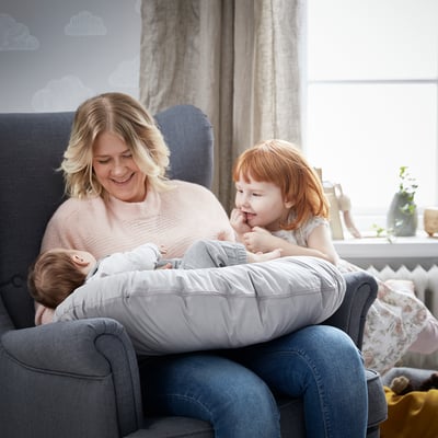 Naine ja kaks last naudivad hubast hetke hallil diivanil koos imetamispadjaga, ümbritsetud pehme sisekujundusega hästi valgustatud toas.