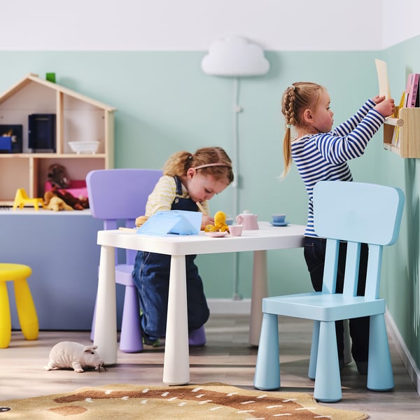 Lapsed mängivad mänguteega värvilise laua taga, üks sinisel toolil, teine lillal taburetil, valge nukumaja lähedal.
