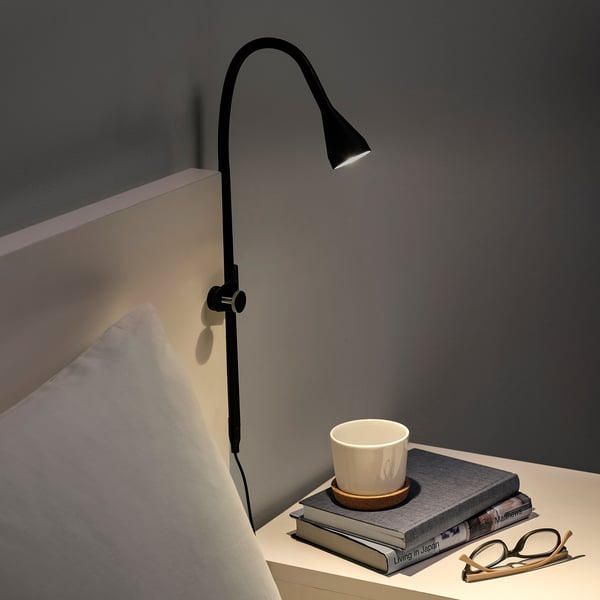 Must reguleeritav klamberlamp voodi kõrval, mis valgustab raamatuvirna, mille peal on kruus.