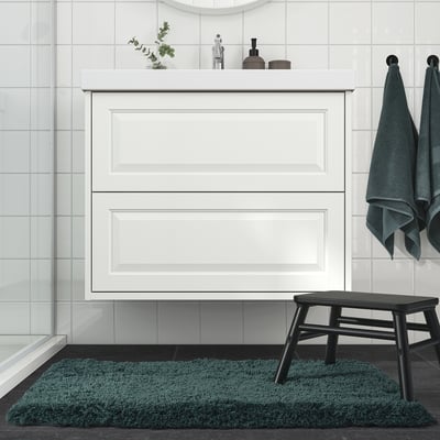 Modern bathroom with ALMTJÄRN teal bath rug, white cabinet, black stool, hanging towels, and glass shower. Rugs fluffy texture, small size.