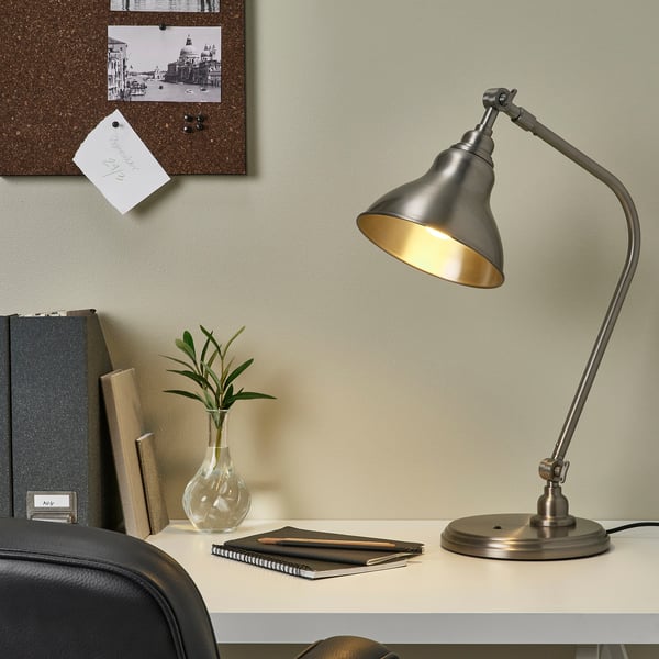 Home office setup with ANKARSPEL lamp. Silver, gooseneck design, bright light. Small vase, books, notebook on desk. Notes on corkboard.