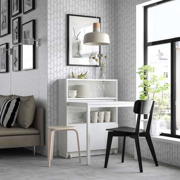 Modern room with BILLY white bookcase desk, black chair, grey sofa, and geometric wallpaper. Large window, black pendant lamp, and framed photos.