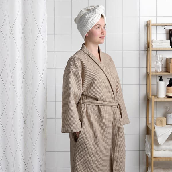 Person in bathrobe with towel turban. Bathrobe has adjustable belt; shelves with toiletries beside.