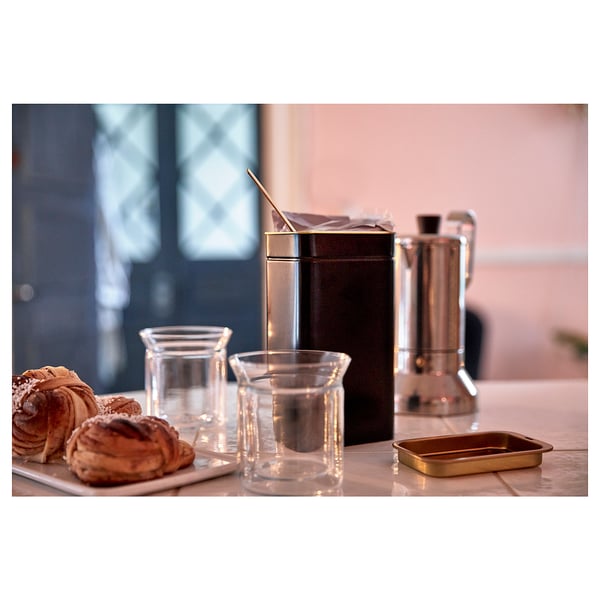 Small copper coffee tin on counter with pastries and glasses. Modern, shiny, cylindrical design.