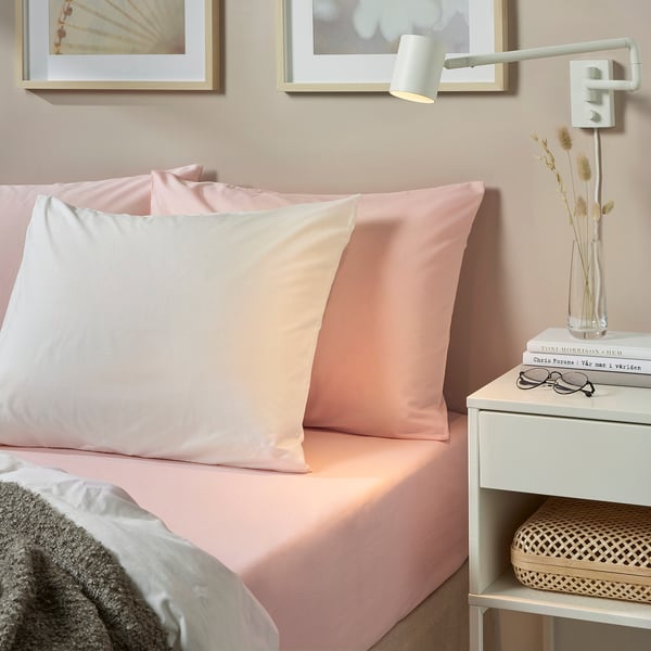 Pastel pink 100% cotton DVALA fitted sheet on bed with pillows, bedside table, and decor.