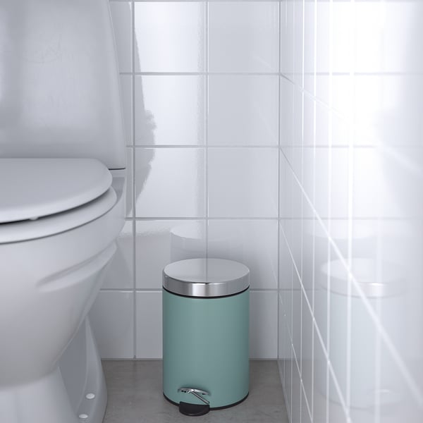 Small green rubbish bin in white tiled bathroom near toilet with foot pedal for hands-free use.