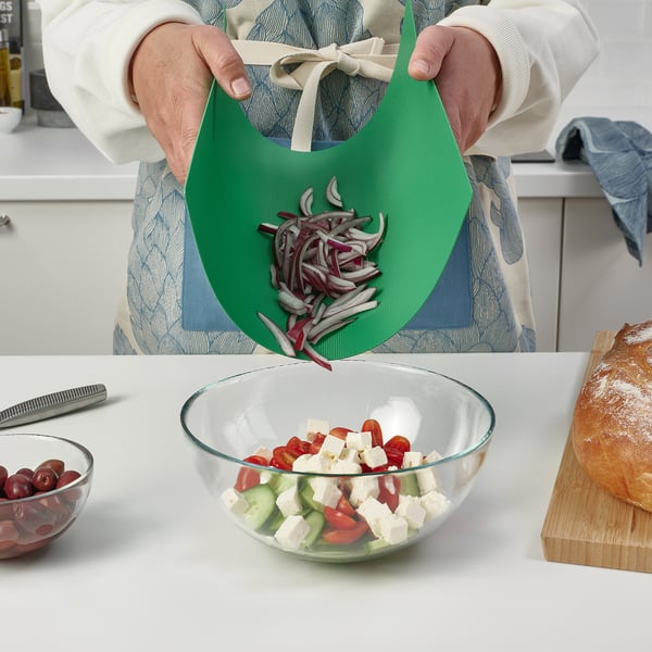 Person holding flexible cutting board with sliced onions, sliding them off into a salad bowl containing tomatoes, cucumbers, and cheese.