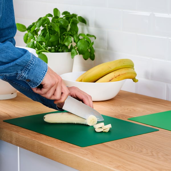 Person slicing banana on flexible green cutting mat, protecting the wooden countertop.
